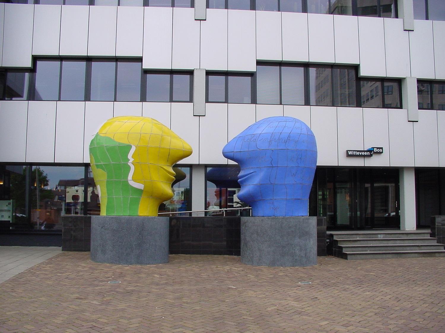 Front view of our headquarters in Deventer. The building is white with black windows and there is an artwork of two large heads in front of it.