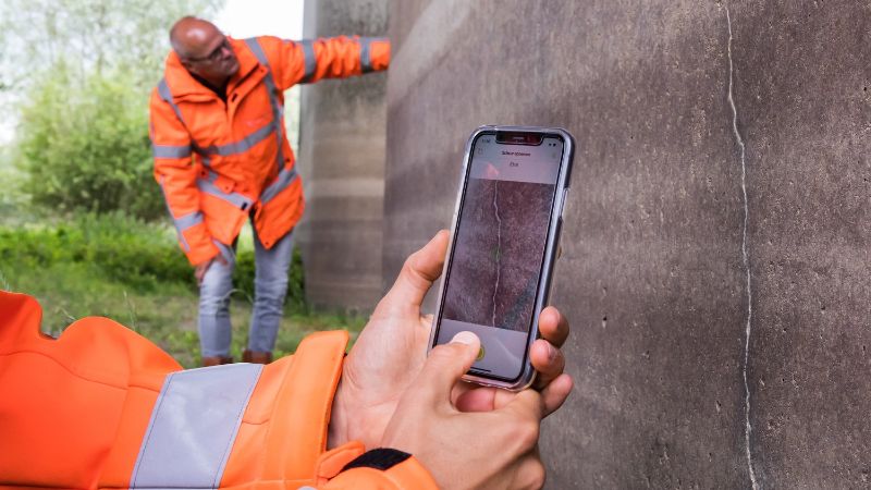 Two hands are holding a phone near a crack in the wall. You can see the Crack Width Analysis app, which is scanning the wall. In the background, another person is checking the wall.