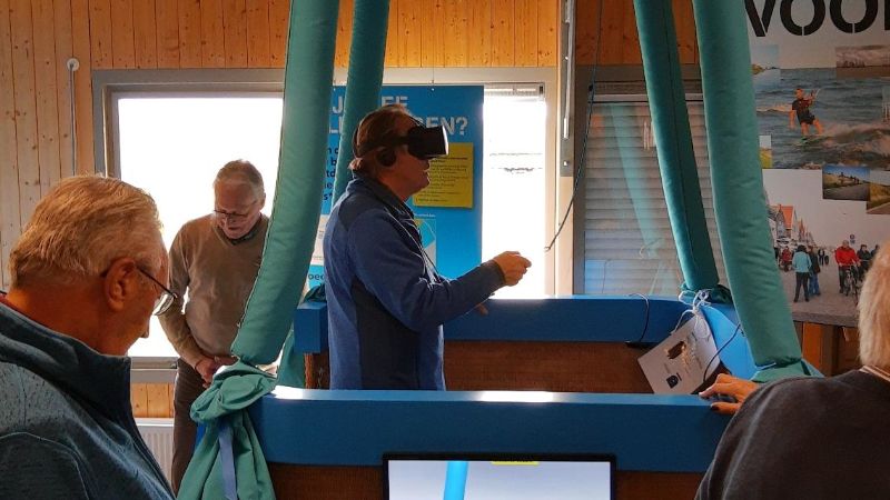A man is wearing VR glasses in the basket of the balloon on the ground. On a television screen you can see how the man is flying above the dyke.
