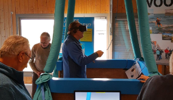 A man is wearing VR glasses in the basket of the balloon on the ground. On a television screen you can see how the man is flying above the dyke.